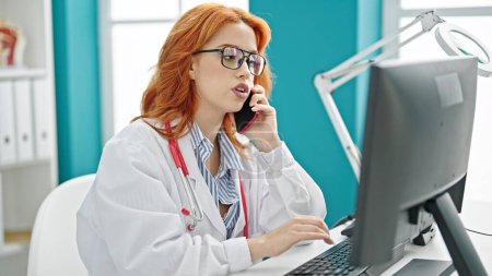 Foto de Mujer pelirroja joven médico usando el ordenador hablando en el teléfono inteligente en la clínica - Imagen libre de derechos
