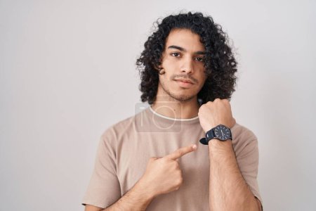 Téléchargez les photos : Homme hispanique avec les cheveux bouclés debout sur fond blanc à la hâte pointant vers regarder le temps, l'impatience, regardant la caméra avec une expression détendue - en image libre de droit