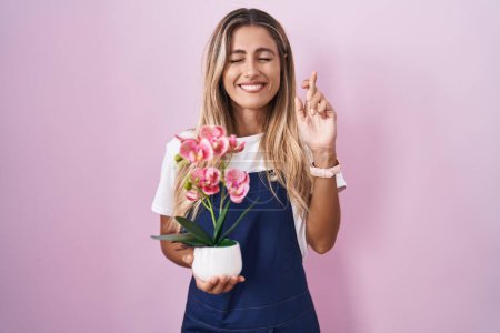 Foto de Young blonde woman wearing gardener apron holding plant gesturing finger crossed smiling with hope and eyes closed. luck and superstitious concept. - Imagen libre de derechos