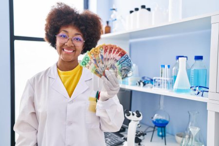 Téléchargez les photos : Jeune femme afro-américaine travaillant au laboratoire scientifique tenant de l'argent regardant positif et heureux debout et souriant avec un sourire confiant montrant les dents - en image libre de droit