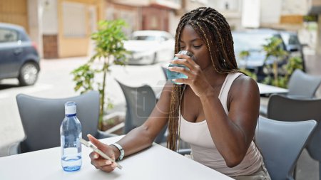 Téléchargez les photos : Femme afro-américaine buvant un verre d'eau en utilisant un smartphone sur la terrasse du café - en image libre de droit