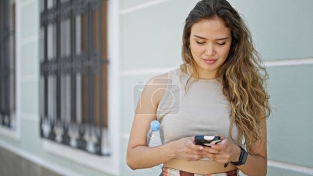 Photo for Young beautiful hispanic woman using smartphone holding bottle of water at street - Royalty Free Image