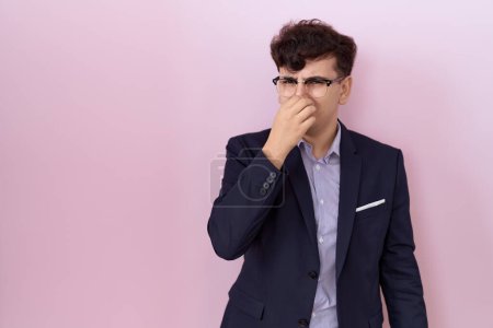 Téléchargez les photos : Jeune homme non binaire avec barbe en costume et cravate sentant quelque chose de puant et dégoûtant, odeur intolérable, retenant son souffle avec les doigts sur le nez. mauvaise odeur - en image libre de droit