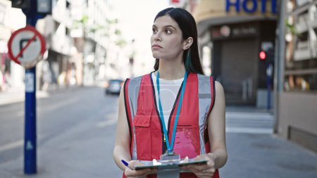 Téléchargez les photos : Jeune belle hispanique femme sondage intervieweur tenant presse-papiers debout avec une expression sérieuse à la rue - en image libre de droit