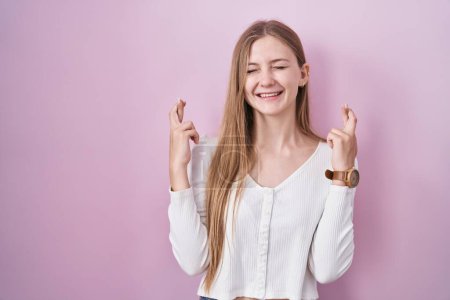 Téléchargez les photos : Jeune femme caucasienne debout sur fond rose geste doigt croisé souriant avec espoir et les yeux fermés. chance et concept superstitieux. - en image libre de droit