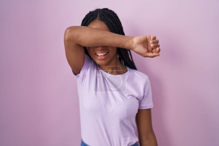 Photo for African american woman with braids standing over pink background covering eyes with arm smiling cheerful and funny. blind concept. - Royalty Free Image