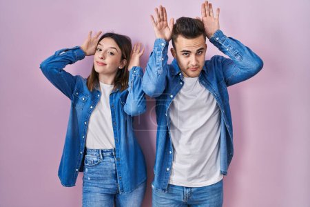 Photo for Young hispanic couple standing over pink background doing bunny ears gesture with hands palms looking cynical and skeptical. easter rabbit concept. - Royalty Free Image