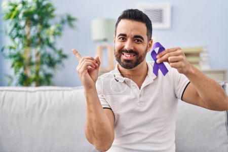 Téléchargez les photos : Jeune homme hispanique avec barbe tenant un ruban violet conscience souriant heureux pointant avec la main et le doigt sur le côté - en image libre de droit