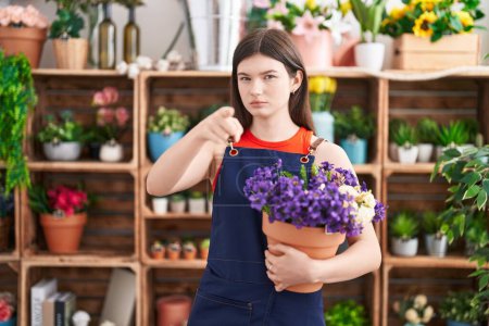 Téléchargez les photos : Jeune femme caucasienne travaillant au magasin de fleuriste tenant pot avec des fleurs pointant du doigt à la caméra et à vous, geste confiant regardant sérieux - en image libre de droit