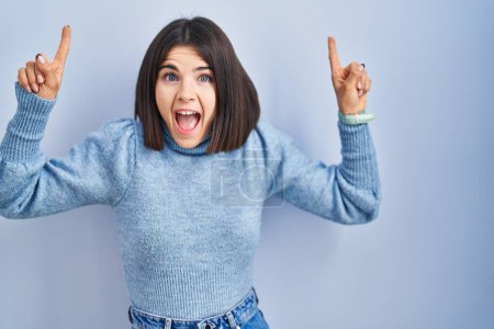 Photo for Young hispanic woman standing over blue background smiling amazed and surprised and pointing up with fingers and raised arms. - Royalty Free Image
