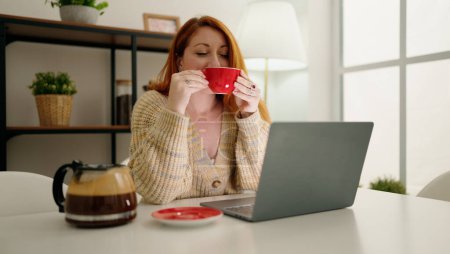 Foto de Joven pelirroja usando laptop bebiendo café en casa - Imagen libre de derechos