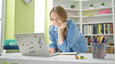 Téléchargez les photos : Jeune étudiante blonde utilisant un ordinateur portable souriant à la bibliothèque de l'université - en image libre de droit