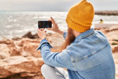 Foto de Joven pelirrojo de pie en la vista trasera hacer foto por teléfono inteligente en la playa - Imagen libre de derechos