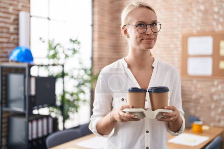 Photo for Young caucasian woman working at the office holding coffee cups smiling looking to the side and staring away thinking. - Royalty Free Image