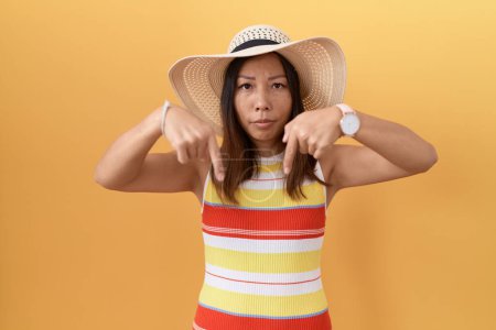 Téléchargez les photos : Femme chinoise du Moyen Âge portant un chapeau d'été sur fond jaune pointant vers le bas regardant triste et contrarié, indiquant la direction avec les doigts, malheureux et déprimé. - en image libre de droit