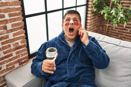 Photo for Hispanic young man wearing bathrobe and eye bags patches drinking wine speaking on the phone angry and mad screaming frustrated and furious, shouting with anger looking up. - Royalty Free Image