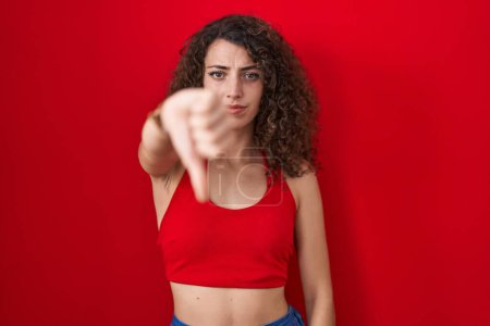 Photo for Hispanic woman with curly hair standing over red background looking unhappy and angry showing rejection and negative with thumbs down gesture. bad expression. - Royalty Free Image