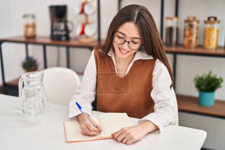 Téléchargez les photos : Jeune belle femme hispanique écrit sur un cahier assis sur la table à la maison - en image libre de droit
