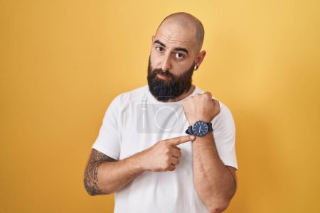 Foto de Joven hombre hispano con barba y tatuajes de pie sobre fondo amarillo con prisa apuntando a ver el tiempo, la impaciencia, mirando a la cámara con expresión relajada - Imagen libre de derechos