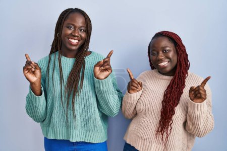 Foto de Dos mujeres africanas de pie sobre fondo azul sonriendo confiadas señalando con los dedos a diferentes direcciones. espacio de copia para publicidad - Imagen libre de derechos