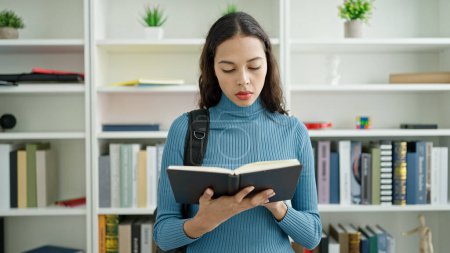 Photo for Young beautiful hispanic woman student standing reading book at university classroom - Royalty Free Image