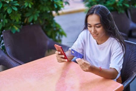 Téléchargez les photos : Young beautiful hispanic woman using smartphone and credit card sitting on table at coffee shop terrace - en image libre de droit