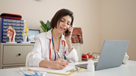 Photo for Young caucasian woman doctor talking on phone writing on document at clinic - Royalty Free Image