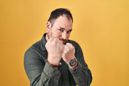 Téléchargez les photos : Plus size hispanic man with beard standing over yellow background ready to fight with fist defense gesture, angry and upset face, afraid of problem - en image libre de droit