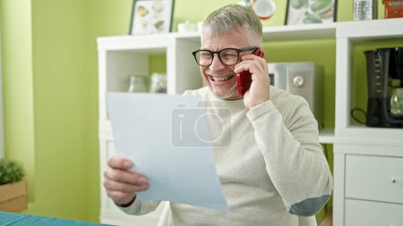 Téléchargez les photos : Moyen-âge homme aux cheveux gris parler sur smartphone document de lecture à la maison - en image libre de droit