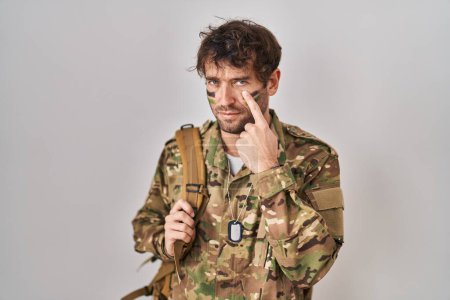 Téléchargez les photos : Jeune homme hispanique portant un uniforme de camouflage de l'armée pointant vers l'œil vous regardant geste, expression suspecte - en image libre de droit