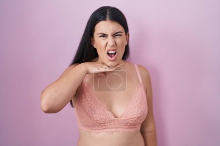 Téléchargez les photos : Jeune femme hispanique portant un soutien-gorge rose tranchant la gorge avec la main comme couteau, menace d'agression avec une violence furieuse - en image libre de droit