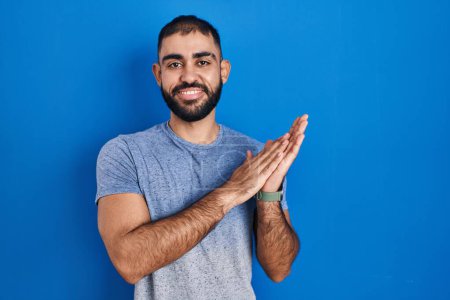 Foto de Hombre de Oriente Medio con barba de pie sobre fondo azul aplaudiendo y aplaudiendo felices y alegres, sonrientes manos orgullosas juntas - Imagen libre de derechos