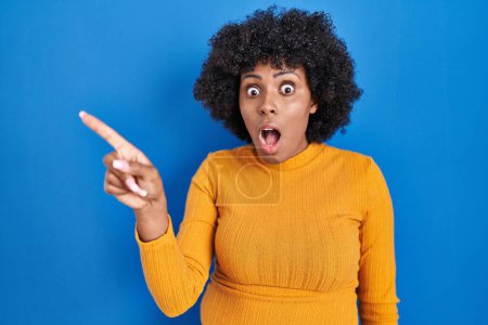 Téléchargez les photos : Femme noire avec des cheveux bouclés debout sur fond bleu surpris pointant du doigt sur le côté, bouche ouverte expression étonnée. - en image libre de droit
