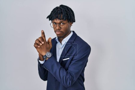 Téléchargez les photos : Jeune homme africain avec dreadlocks portant une veste d'affaires sur fond blanc tenant un pistolet symbolique avec geste de la main, jouant tuer des armes de tir, visage en colère - en image libre de droit