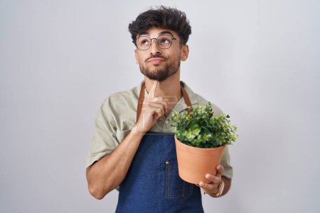 Téléchargez les photos : Homme arabe avec barbe tenant pot de plante verte pensée concentrée sur le doute avec le doigt sur le menton et la recherche jusqu'à se demander - en image libre de droit