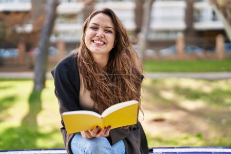 Photo for Young beautiful hispanic woman reading book sitting on bench at park - Royalty Free Image