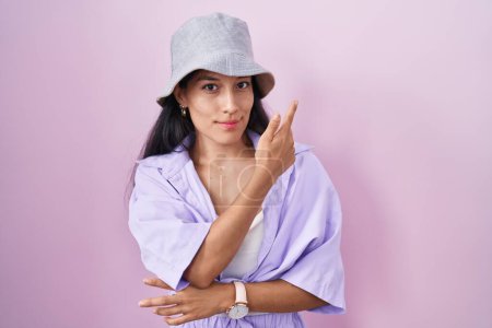 Foto de Mujer hispana joven de pie sobre fondo rosa con sombrero apuntando con el dedo de la mano hacia un lado mostrando publicidad, cara seria y tranquila - Imagen libre de derechos