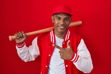 Téléchargez les photos : Jeune homme hispanique jouant au baseball tenant batte gaie avec un sourire sur le visage pointant avec la main et le doigt sur le côté avec une expression heureuse et naturelle - en image libre de droit