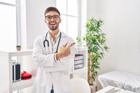 Téléchargez les photos : Homme hispanique portant uniforme de médecin et stéthoscope souriant pointant joyeusement avec la main et le doigt sur le côté - en image libre de droit