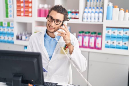 Téléchargez les photos : Jeune homme hispanique pharmacien parlant au téléphone en utilisant l'ordinateur à la pharmacie - en image libre de droit