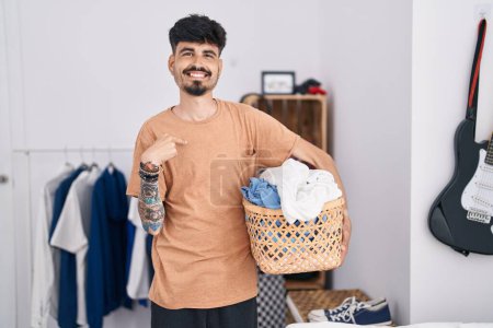 Foto de Joven hombre hispano con barba sosteniendo la cesta de la ropa en el dormitorio señalando con el dedo a uno mismo sonriendo feliz y orgulloso - Imagen libre de derechos