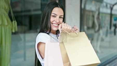 Téléchargez les photos : Jeune belle femme hispanique souriant aller faire du shopping tenant des sacs au magasin de vêtements - en image libre de droit