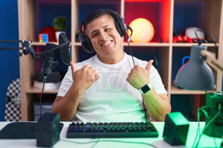 Photo for Young hispanic man playing video games success sign doing positive gesture with hand, thumbs up smiling and happy. cheerful expression and winner gesture. - Royalty Free Image