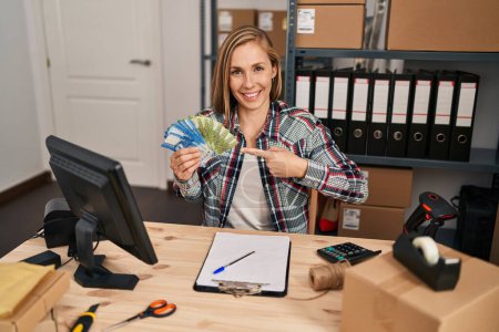 Téléchargez les photos : Jeune femme blonde travaillant dans une petite entreprise e-commerce tenant des billets de banque en pesos chiliens souriant heureux pointant avec la main et le doigt - en image libre de droit