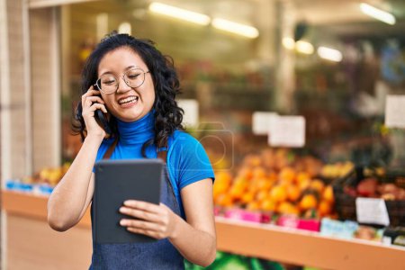 Téléchargez les photos : Young chinese woman employee talking on the smartphone using touchpad at fruit store - en image libre de droit