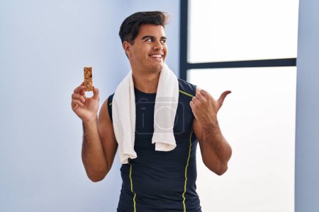 Foto de Hispanic man eating protein bar as healthy energy snack pointing thumb up to the side smiling happy with open mouth - Imagen libre de derechos