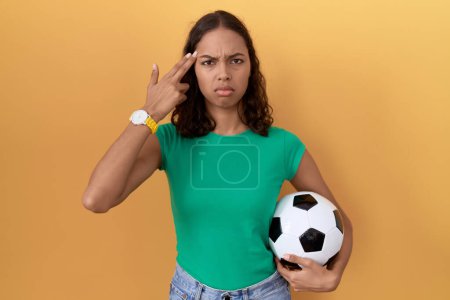 Foto de Joven mujer hispana sosteniendo la pelota disparando y matándose apuntando de la mano y los dedos a la cabeza como arma, gesto suicida. - Imagen libre de derechos