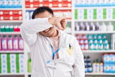 Foto de Joven chino trabajando en farmacia cubriendo los ojos con el brazo sonriendo alegre y divertido. concepto ciego. - Imagen libre de derechos