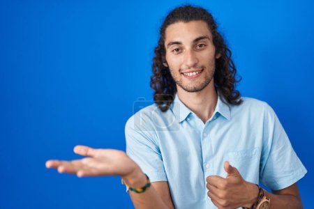 Téléchargez les photos : Young hispanic man standing over blue background showing palm hand and doing ok gesture with thumbs up, smiling happy and cheerful - en image libre de droit