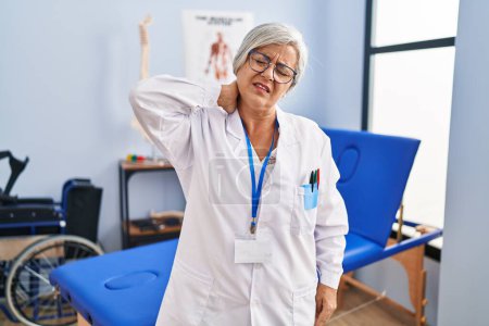 Foto de Mujer de mediana edad con pelo gris que trabaja en la clínica de recuperación del dolor que sufre de dolor de cuello lesión, tocando el cuello con la mano, dolor muscular - Imagen libre de derechos